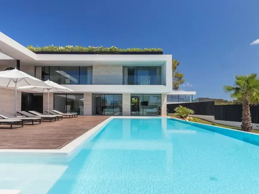 Villa Can Cana en Ibiza vista desde la piscina: dos plantas con revestimiento de travertino, tarima de madera, tumbonas y sombrillas blancas.