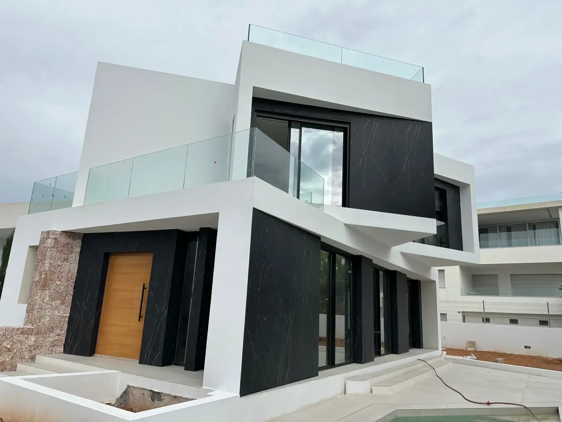 Acceso de una vivienda de obra nueva con puerta de madera, zócalo de piedra y volumen blanco.