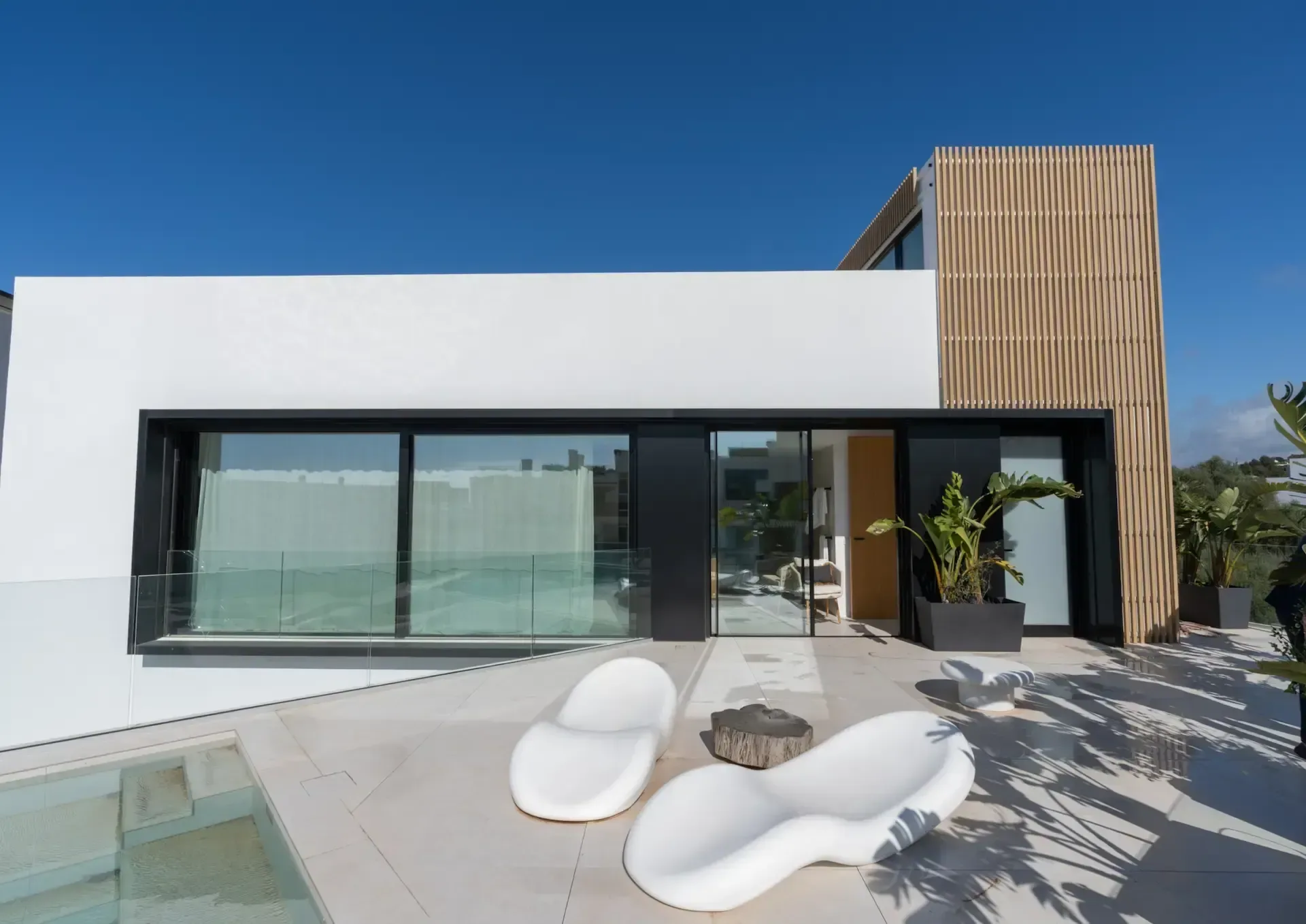 Terraza de una villa construida por Unero en Ibiza, con tumbonas blancas, borde de piscina y celosía vertical de madera bajo un cielo azul intenso.
