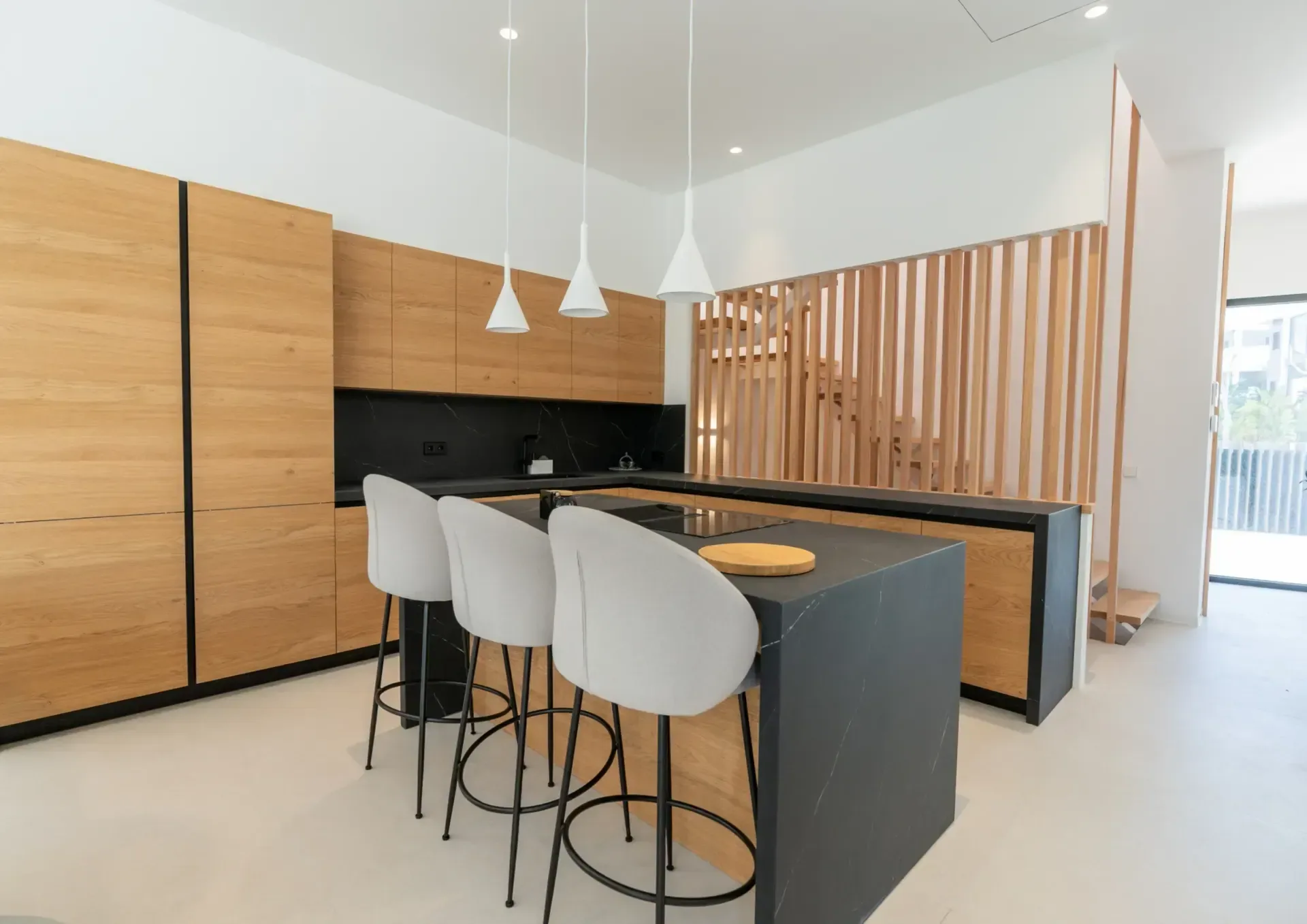Cocina abierta con taburetes altos, columna de armarios de roble hasta el techo y lamado vertical de madera junto a la escalera.
