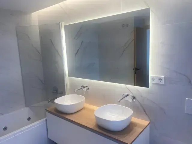 Baño con espejo retroiluminado, dos lavabos sobre encimera continua y mueble suspendido de madera.