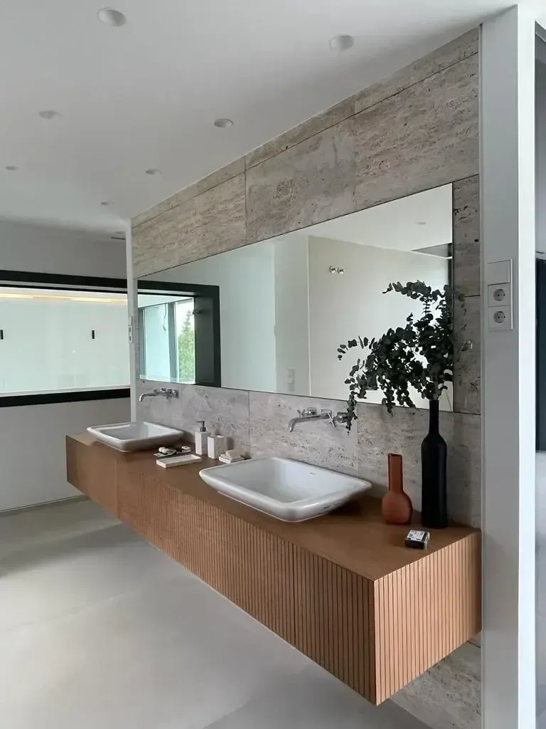 Baño con paño de travertino, dos lavabos sobre encimera y mueble suspendido de madera.