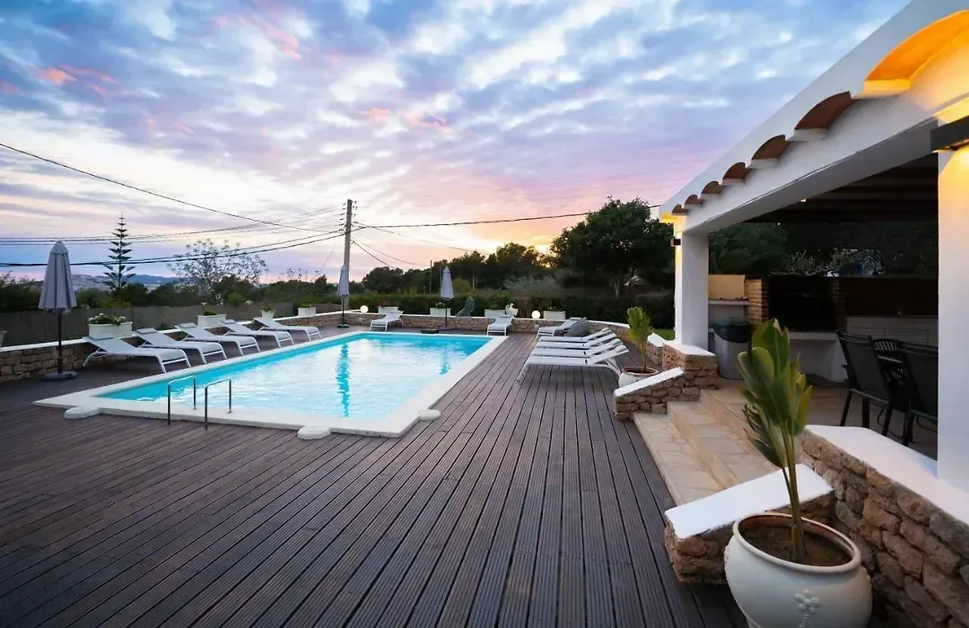 Piscina de Can Flowers al atardecer, con tarima de madera, tumbonas, macetones plantados y el cielo naranja sobre la casa encalada.