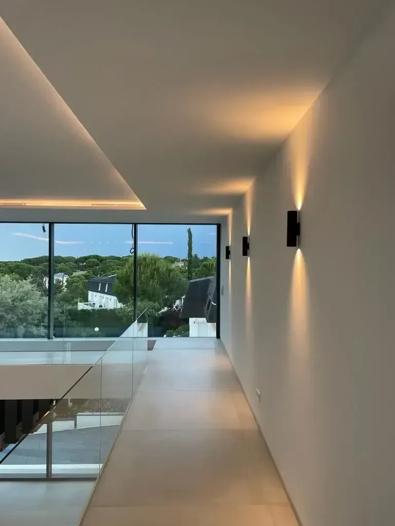 Pasillo con cornisa de luz oculta que lava el techo y ventanal al fondo abierto a la terraza.