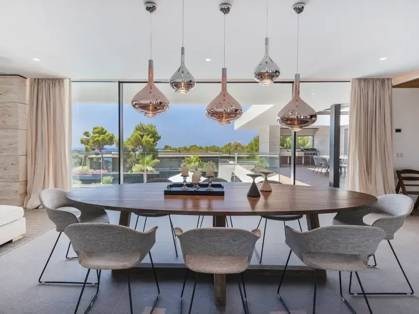 Comedor con mesa larga, lámparas circulares de acabado bronce y ventanal corrido hacia la terraza y las palmeras.