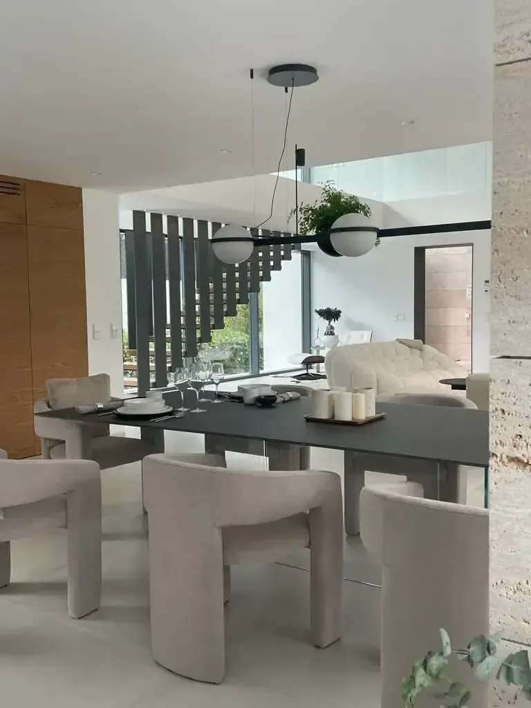 Comedor con sillas escultóricas claras, lámpara negra suspendida y pared de piedra al fondo.