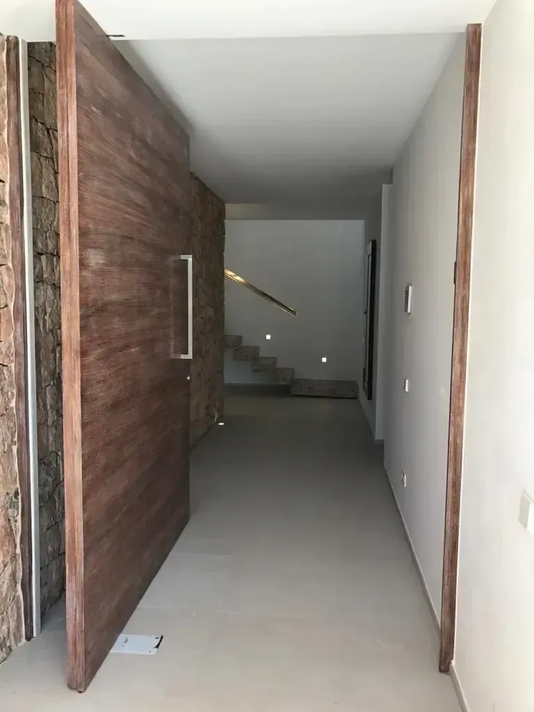 Puerta de acceso pivotante de madera de gran formato, abierta hacia un pasillo de paramentos claros.