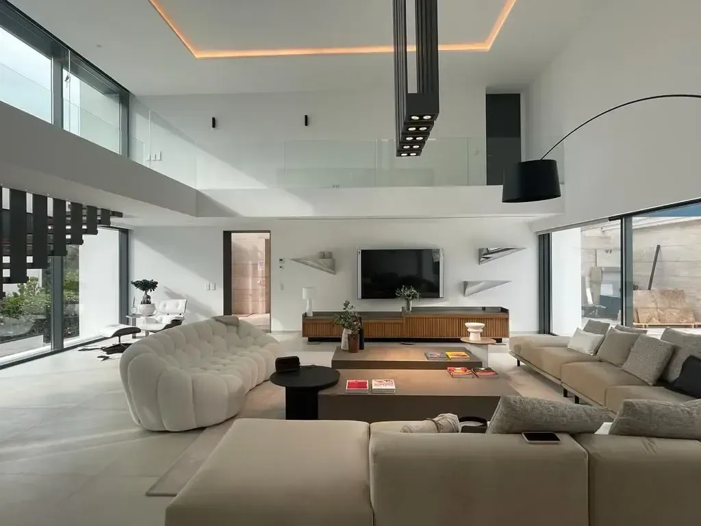 Salón de la vivienda Alina con sofá modular claro, cornisa perimetral iluminada en el techo y frente acristalado al jardín.
