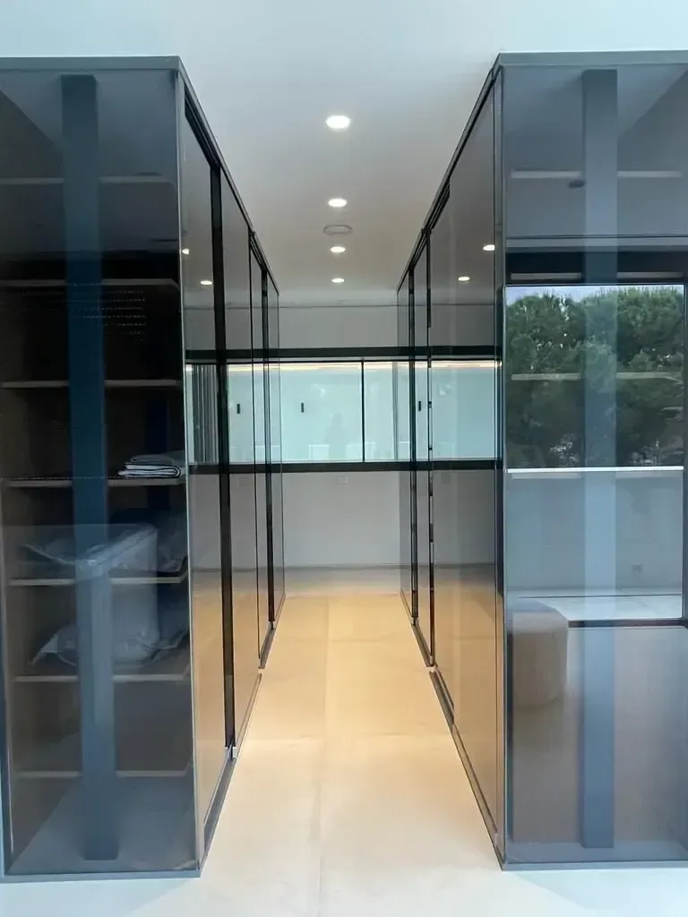 Vestidor con particiones de vidrio y perfilería negra, iluminado con línea continua en el techo.