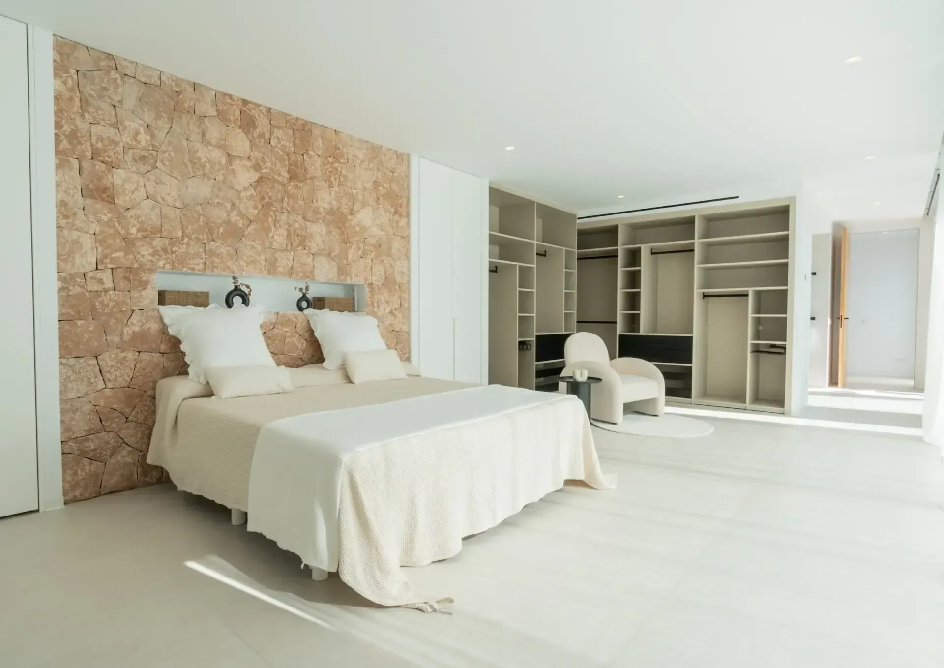 Dormitorio principal con pared de piedra natural tras el cabecero y paso abierto al vestidor.