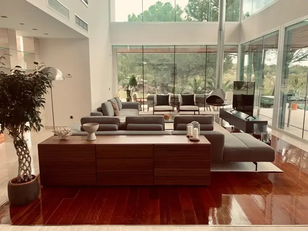 Salón a doble altura con sofás claros, suelo de madera oscura y ventanales al jardín.