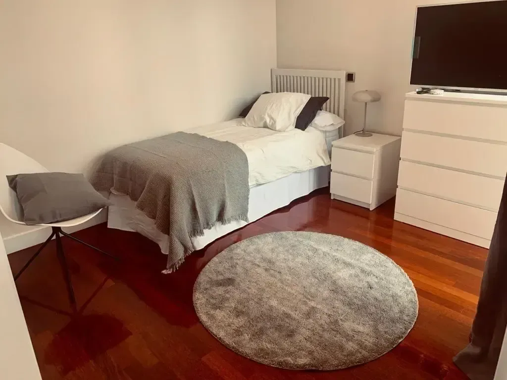 Dormitorio individual con cómoda blanca, alfombra redonda y suelo de madera.