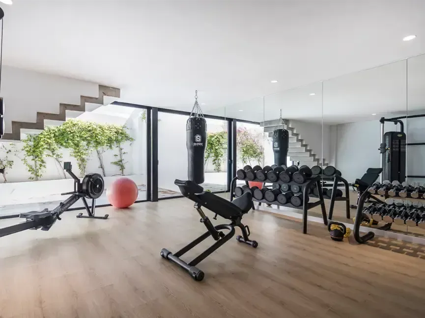 Gimnasio con máquinas de entrenamiento, suelo de madera y luz natural desde el patio.